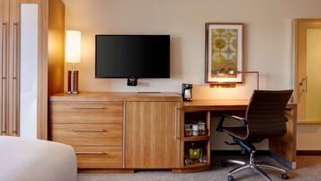 A hotel room with a wooden desk, a flat-screen TV, a lamp, a framed artwork, and a chair.