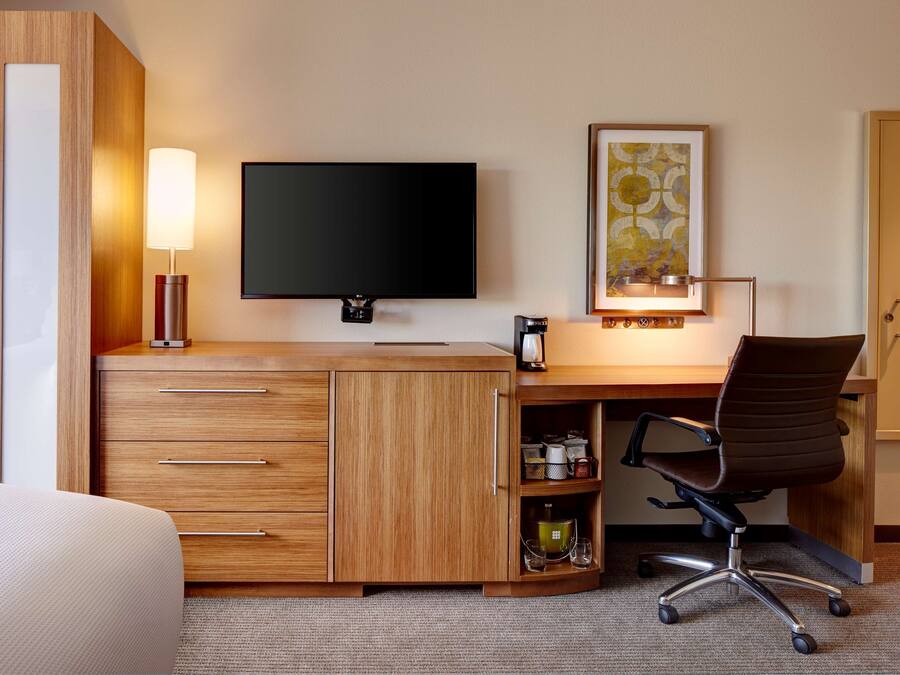 A hotel room with a wooden desk, a flat-screen TV, a lamp, a framed artwork, and a chair.