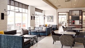 A spacious lounge area with seating and tables, large windows, and a kitchen area visible in the background. - Hyatt Place Madison / Verona (Verona)