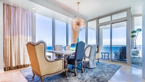 A spacious dining area with a large table and multiple chairs, a modern chandelier, and large windows offering a view of the ocean.