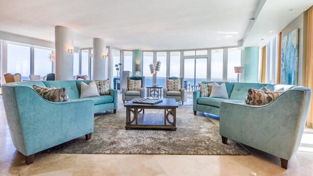 A spacious living area with teal sofas, a central coffee table, and large windows offering a view of the ocean.