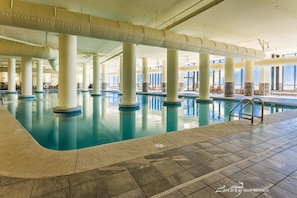 An indoor swimming pool with clear blue water, supported by large white columns, and a tiled floor.