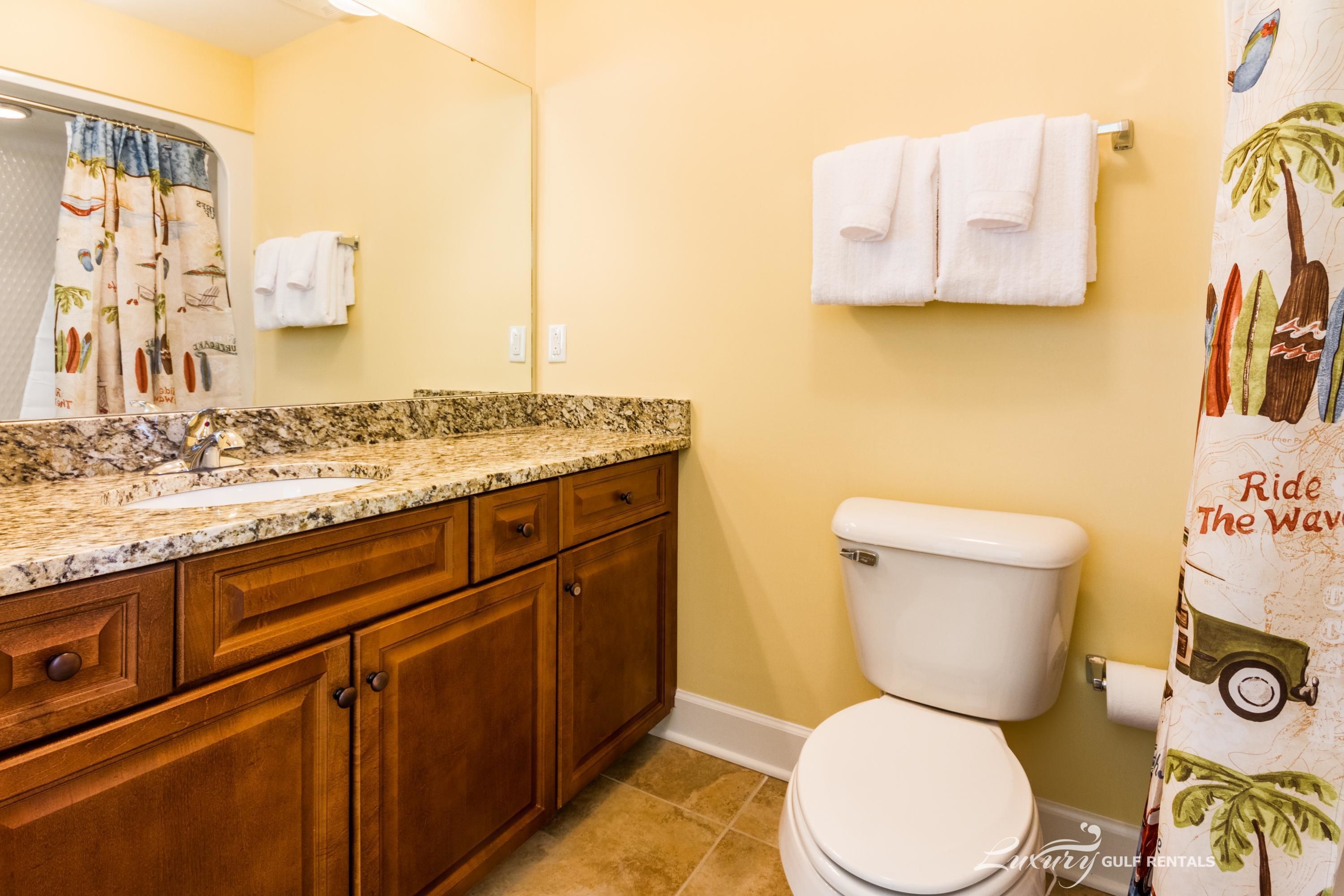 Un baño con lavamanos, armarios de madera, inodoro y cortina de ducha con diseño tropical.