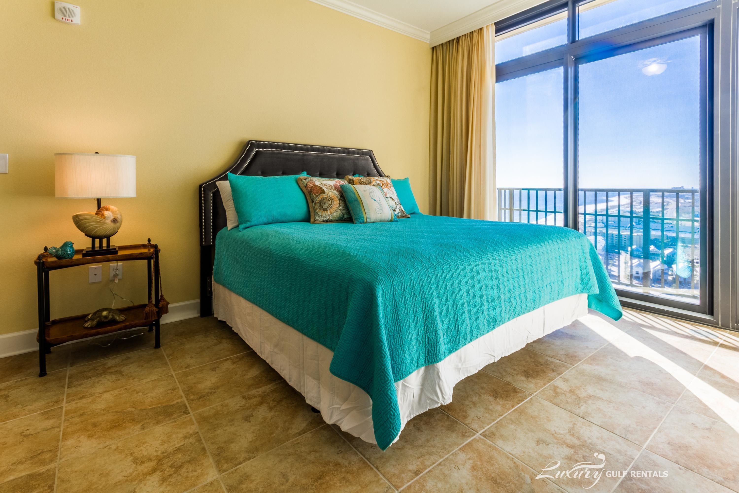 A hotel room with a large bed, a nightstand, a lamp, and a view of the city through large windows.