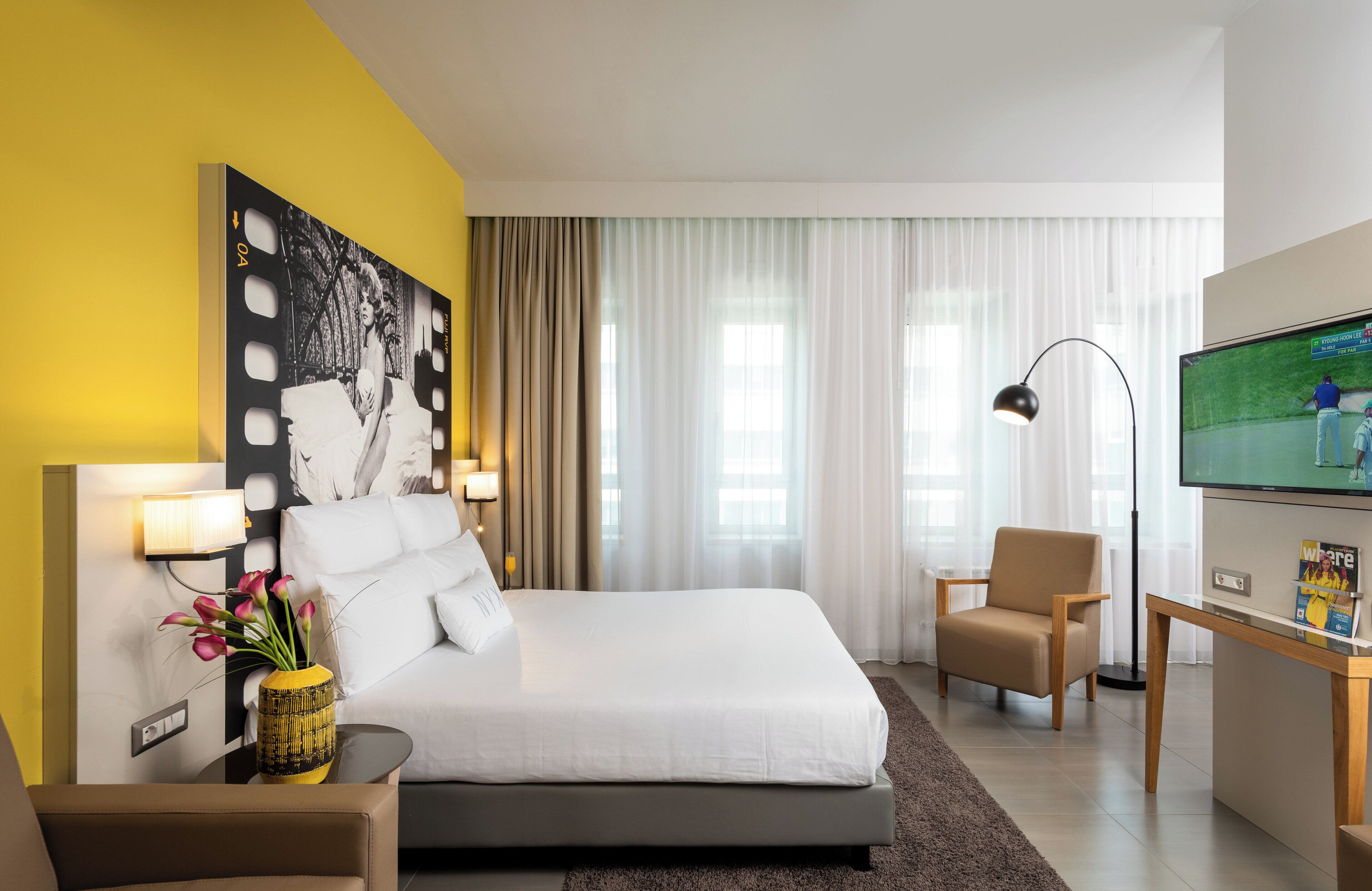 A modern hotel room with a large bed, a desk with a flat-screen TV, a chair, and a vase with flowers.