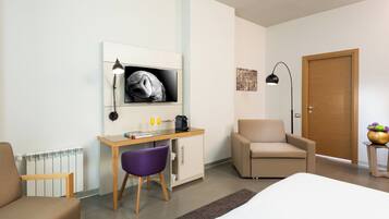 A hotel room with a bed, a desk with a computer, a chair, a sofa, a door, and a small table with a plant.