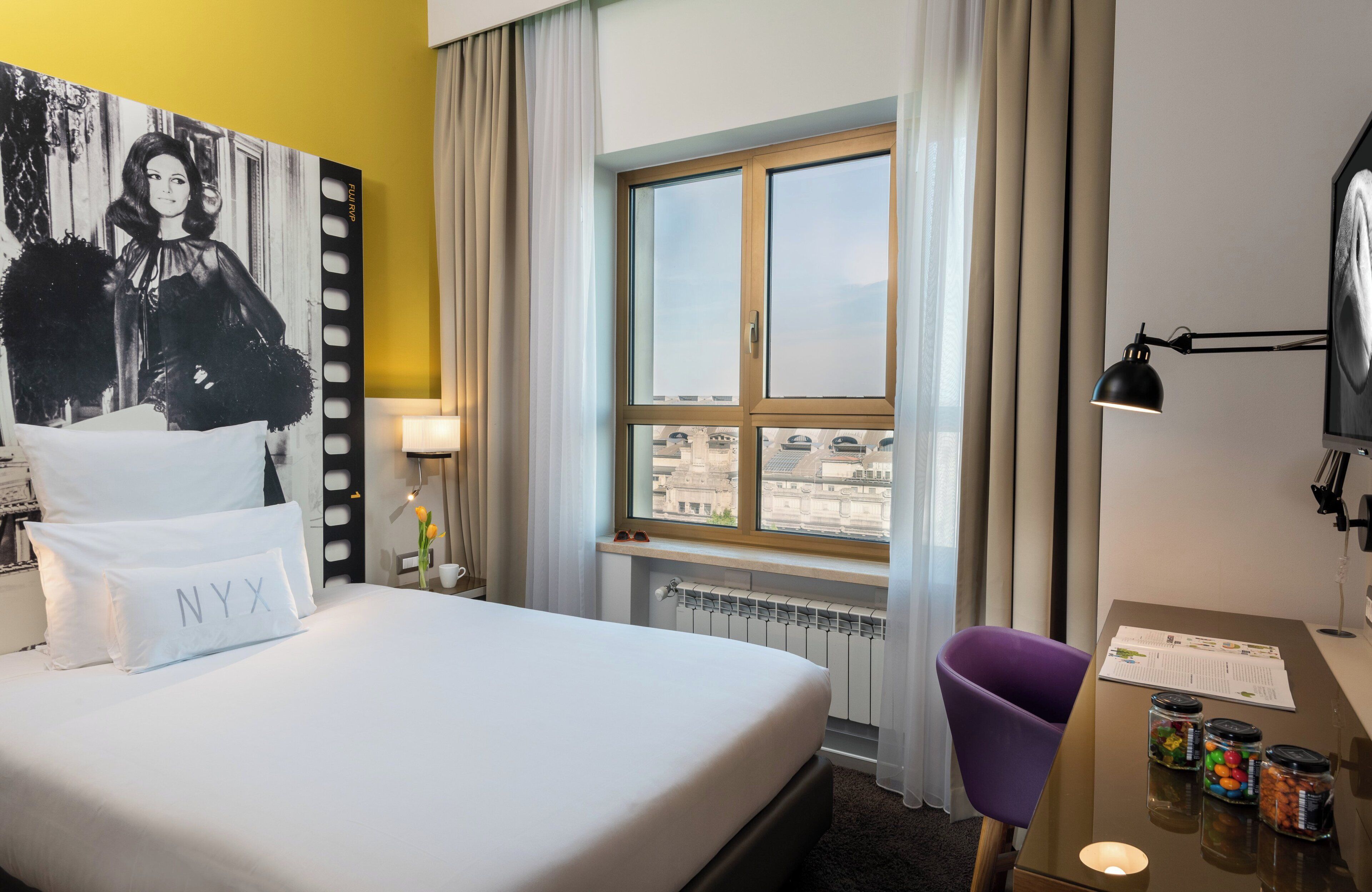 Une chambre d’hôtel avec un lit, un bureau, une chaise, une fenêtre avec des rideaux et une vue sur la ville.