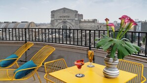Una mesa amarilla con un florero a rayas y una bebida, dos sillas amarillas con cojines azules, y una vista de un edificio histórico al fondo.