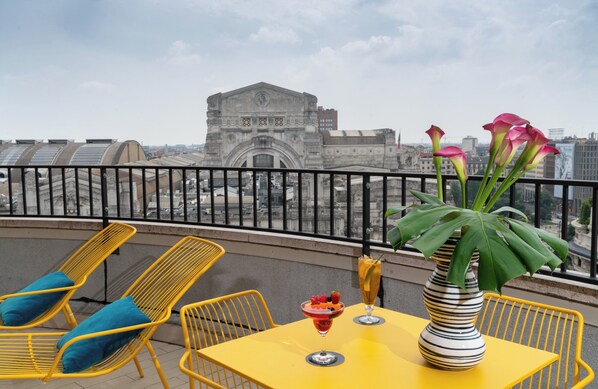A yellow table with a striped vase and a drink, two yellow chairs with blue cushions, and a view of a historic building in the background. - NYX Hotel Milan by Leonardo Hotels (Milan)