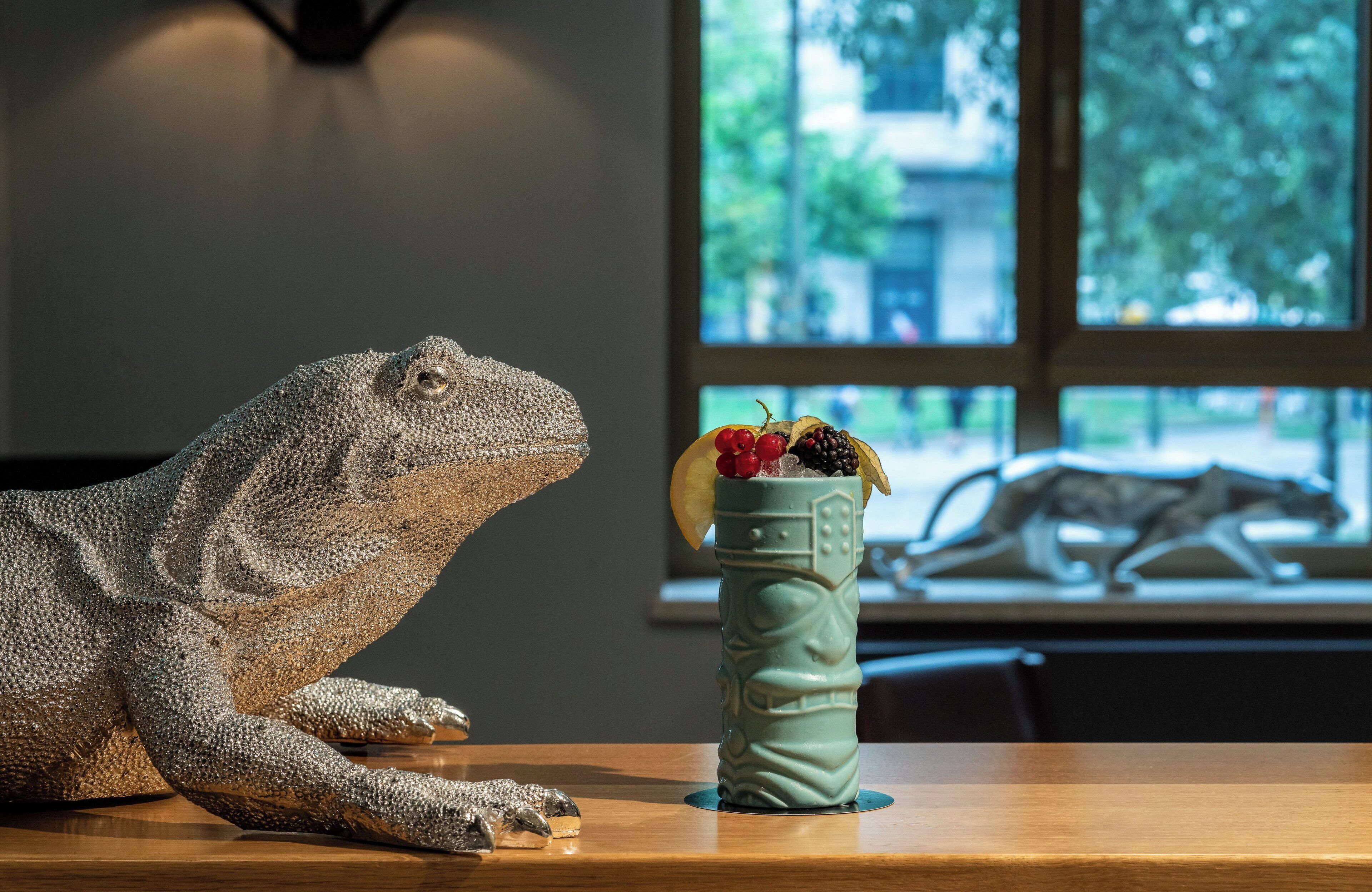 Una escultura de lagarto sobre una mesa de madera con un jarrón decorativo que contiene frutas y una bebida.