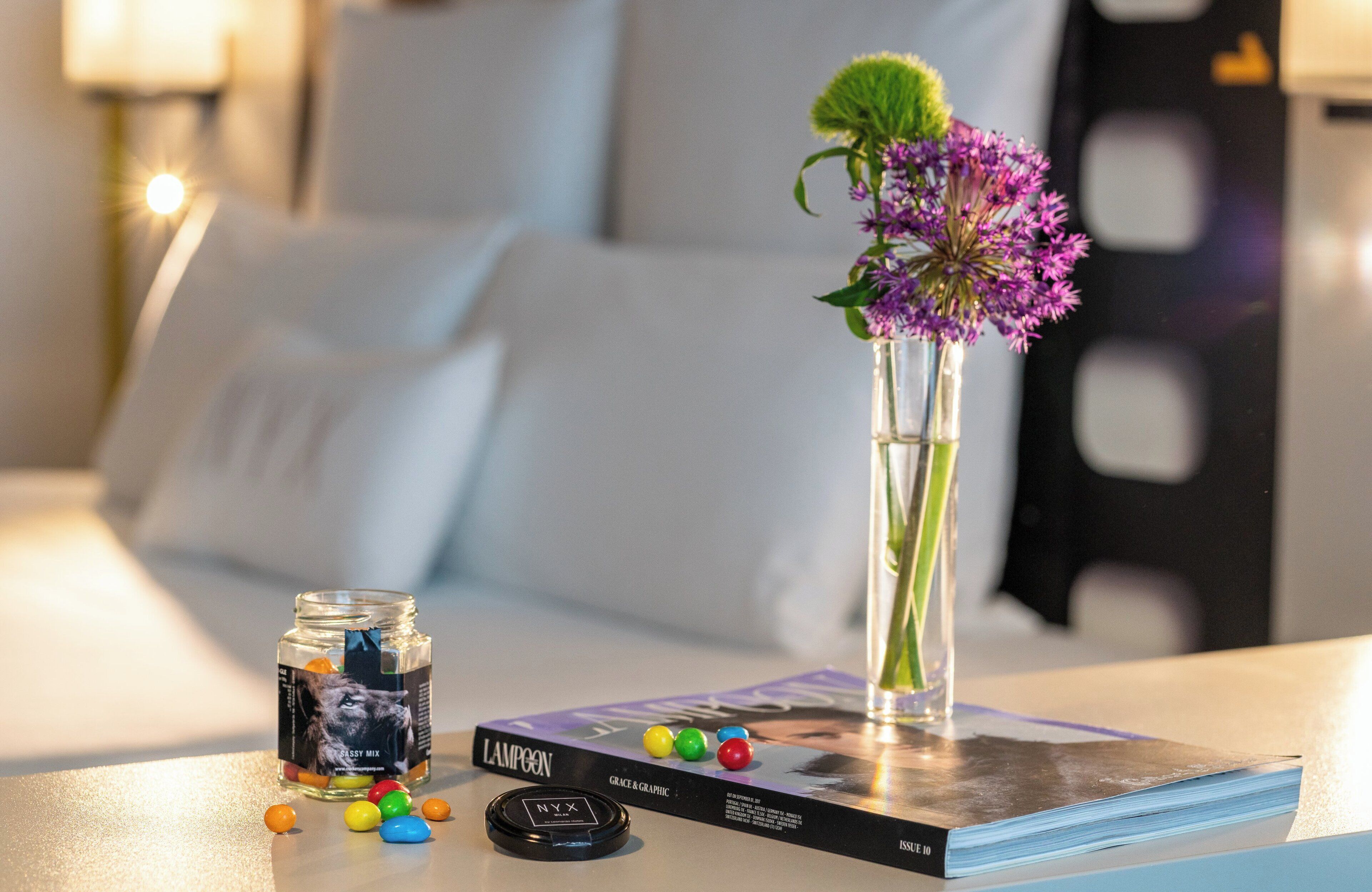 A neatly arranged hotel room with a bed, a bedside table with a book, a vase with flowers, and a jar of candies.