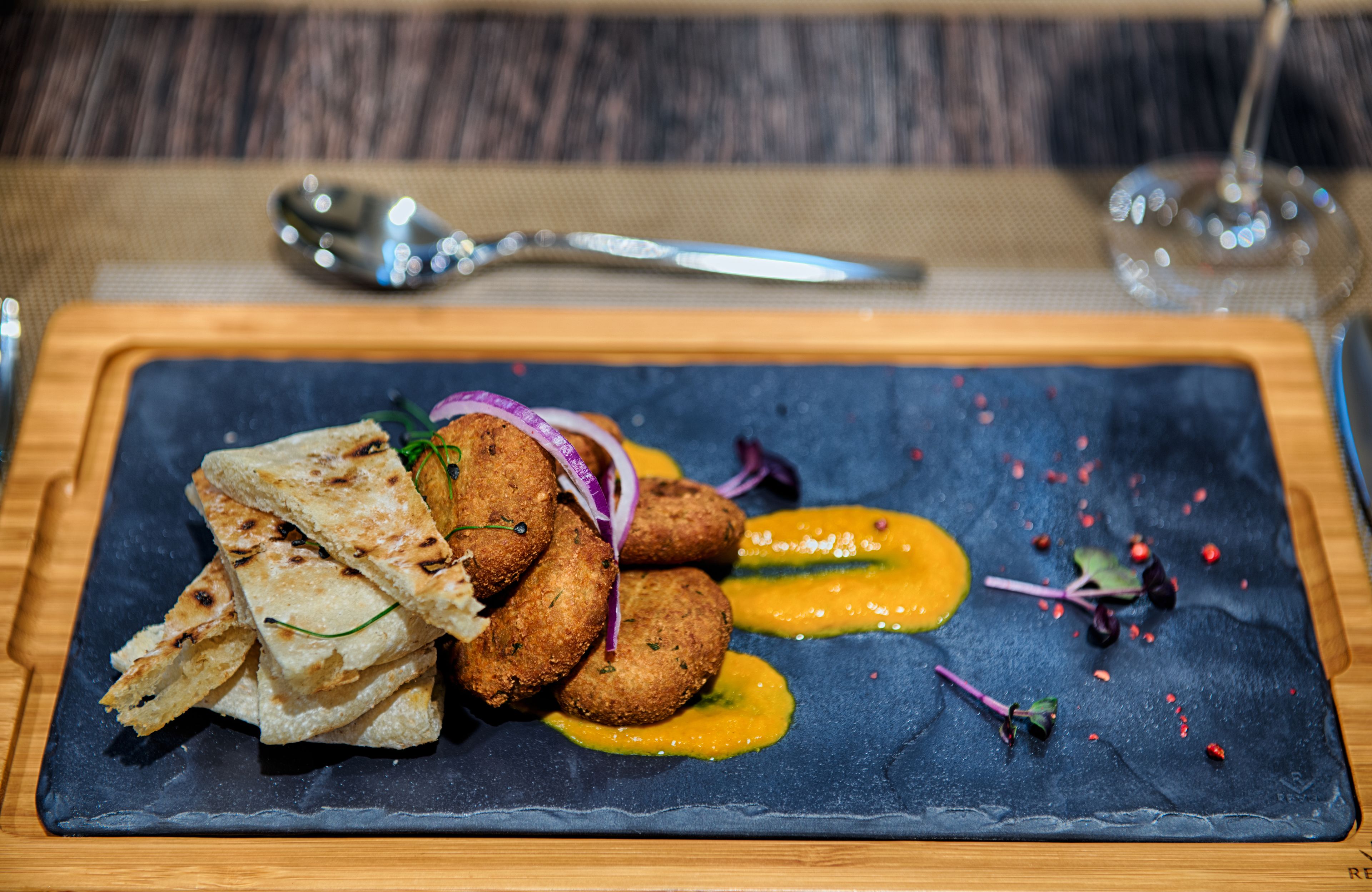 Una tabla de pizarra con comida, una cuchara y una copa de vino sobre una mesa de madera.