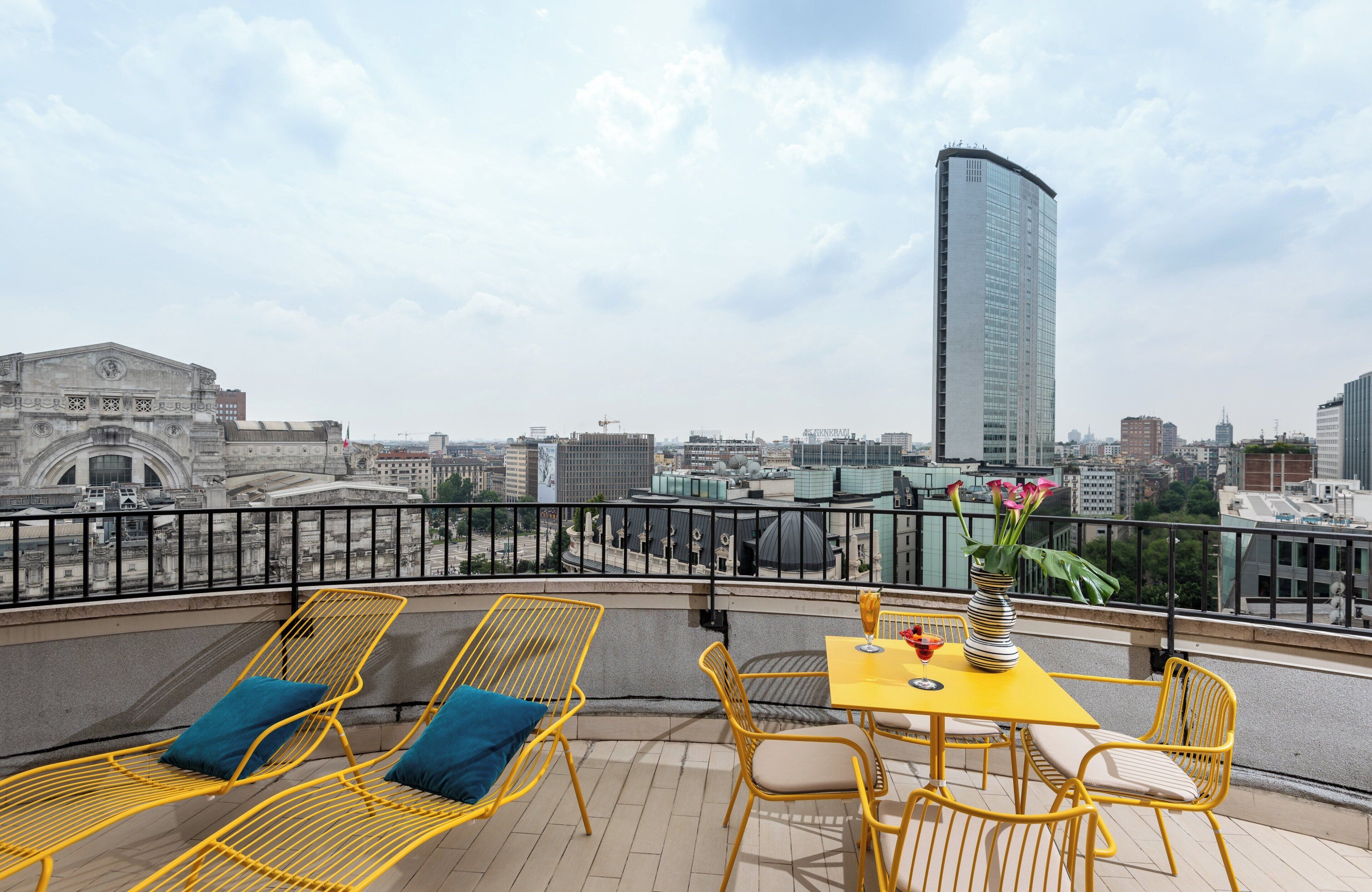 A rooftop terrace with yellow metal chairs and a table, overlooking a cityscape with modern buildings and a historic archway.