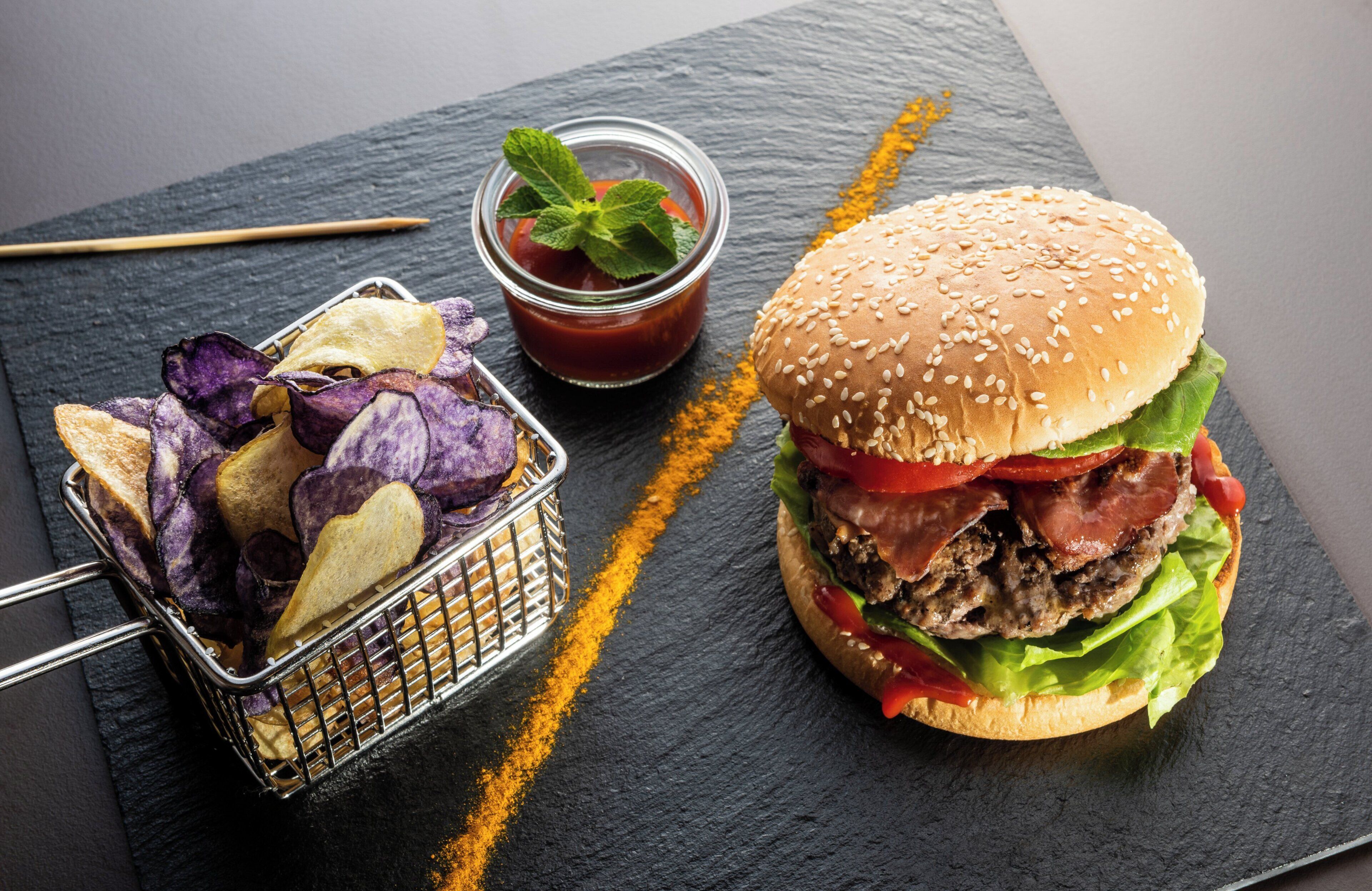 Un hamburger avec de la laitue, des tomates et du bacon, un petit bol de ketchup et un panier de chips, le tout présenté sur une planche en ardoise.