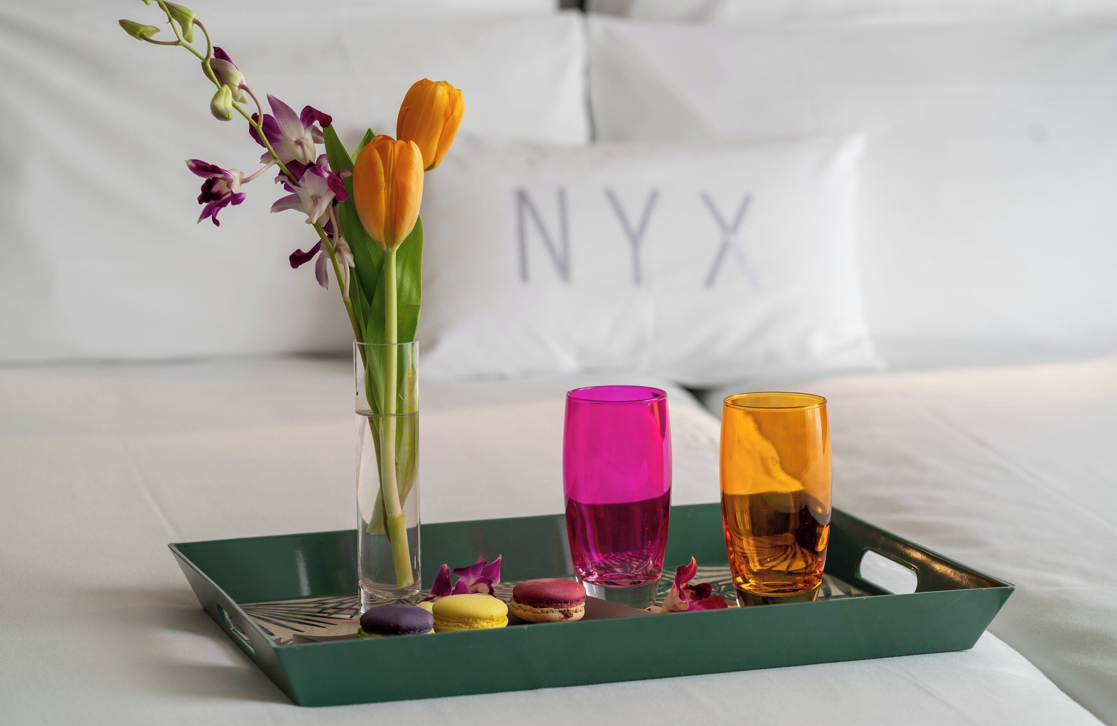 A tray with a vase of flowers, two glasses, and macarons on a bed with a pillow labeled "NYX".