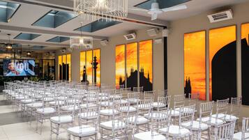 Un vaste hall avec des rangées de chaises et de tables blanches, de grandes fenêtres et des œuvres d’art représentant des paysages urbains.