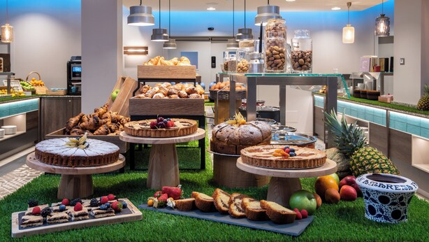 Montaje de bufé con diversas pastas y frutas sobre puestos de madera, una máquina de café y un refrigerador al fondo.