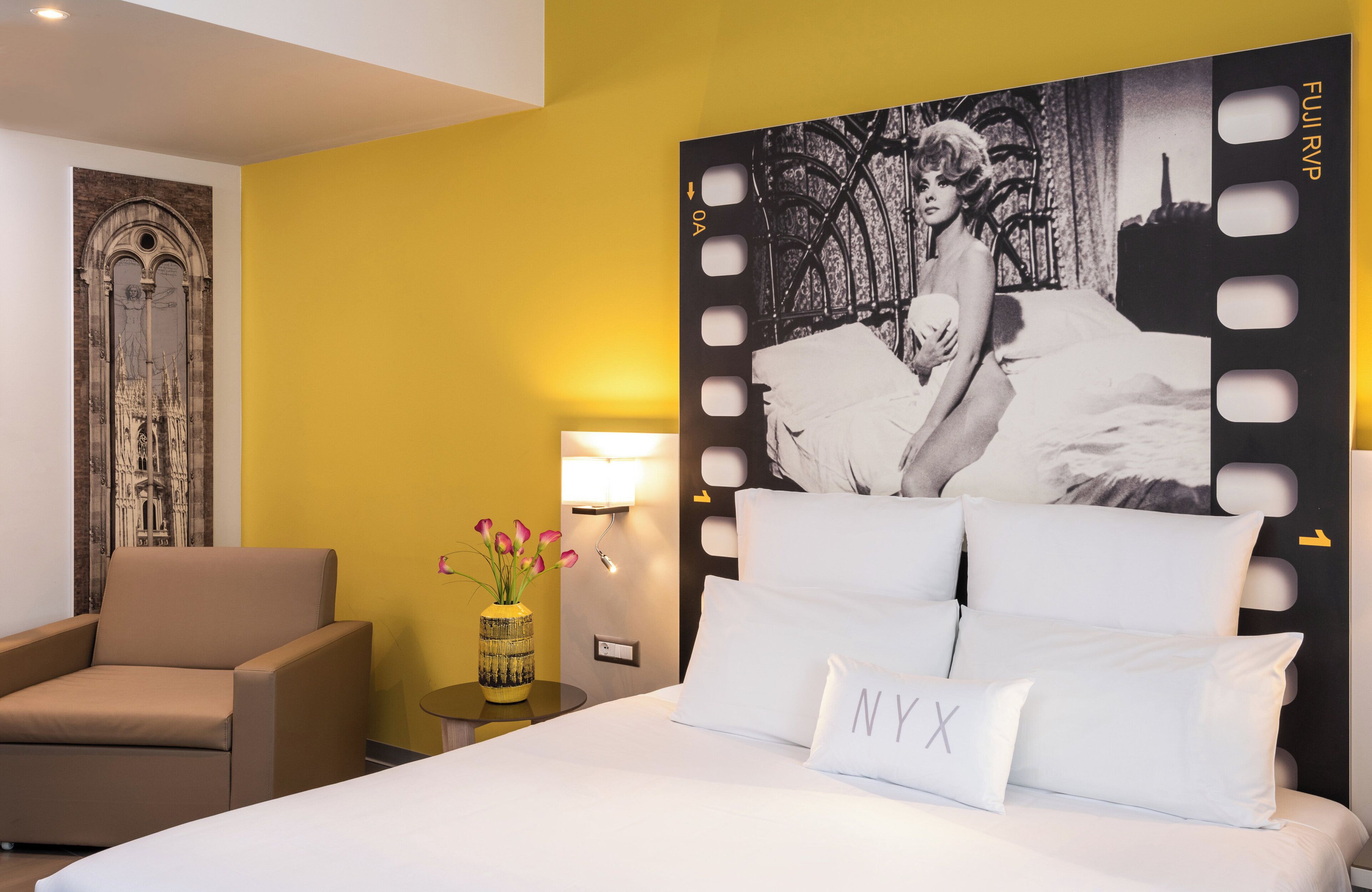 Una habitación de hotel con cama, una silla, una mesita de noche, una lámpara y un cartel grande en la pared.