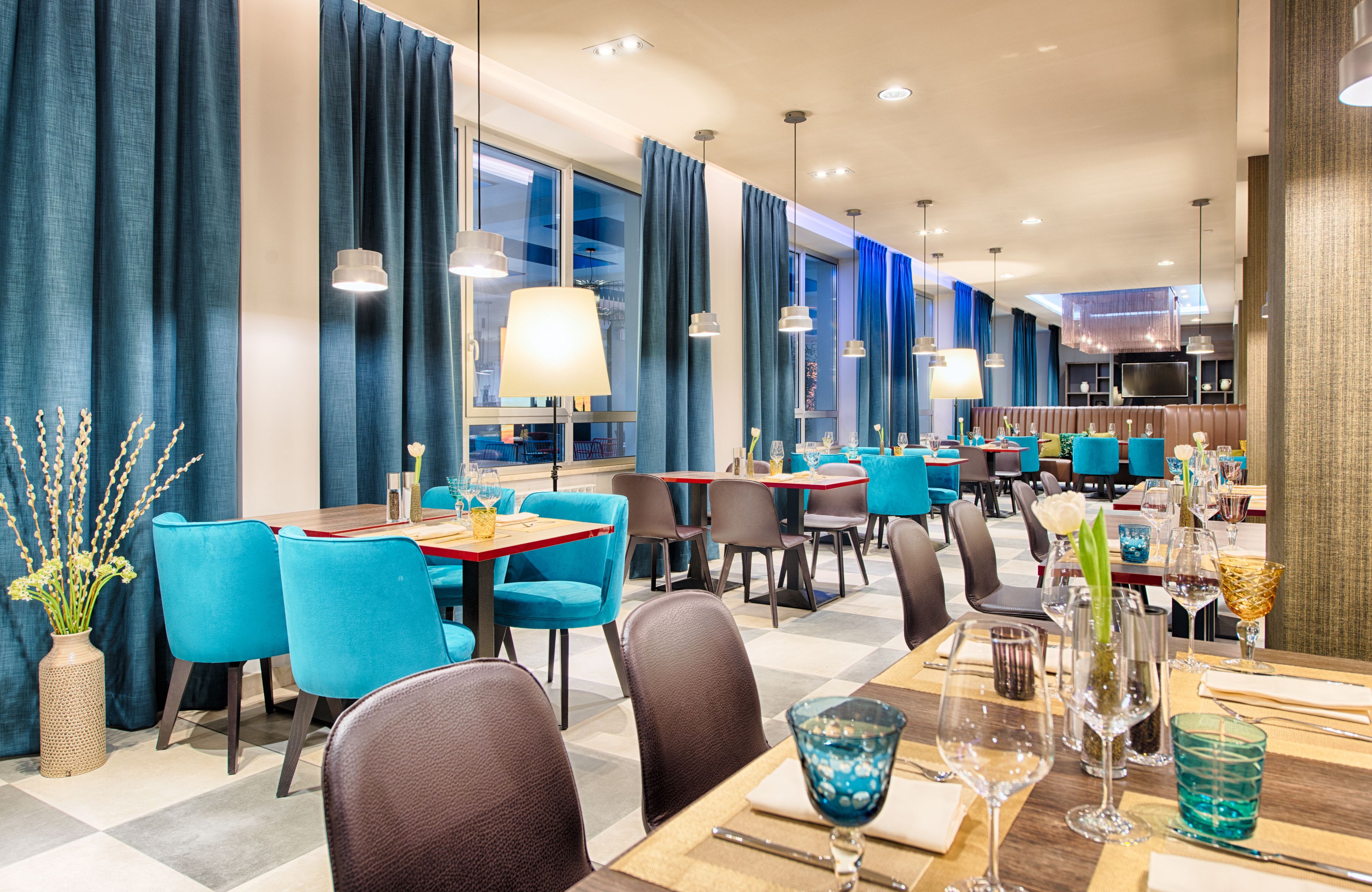 Un comedor de hotel moderno con sillas azules, mesas dispuestas para comer y amplias ventanas con cortinas.