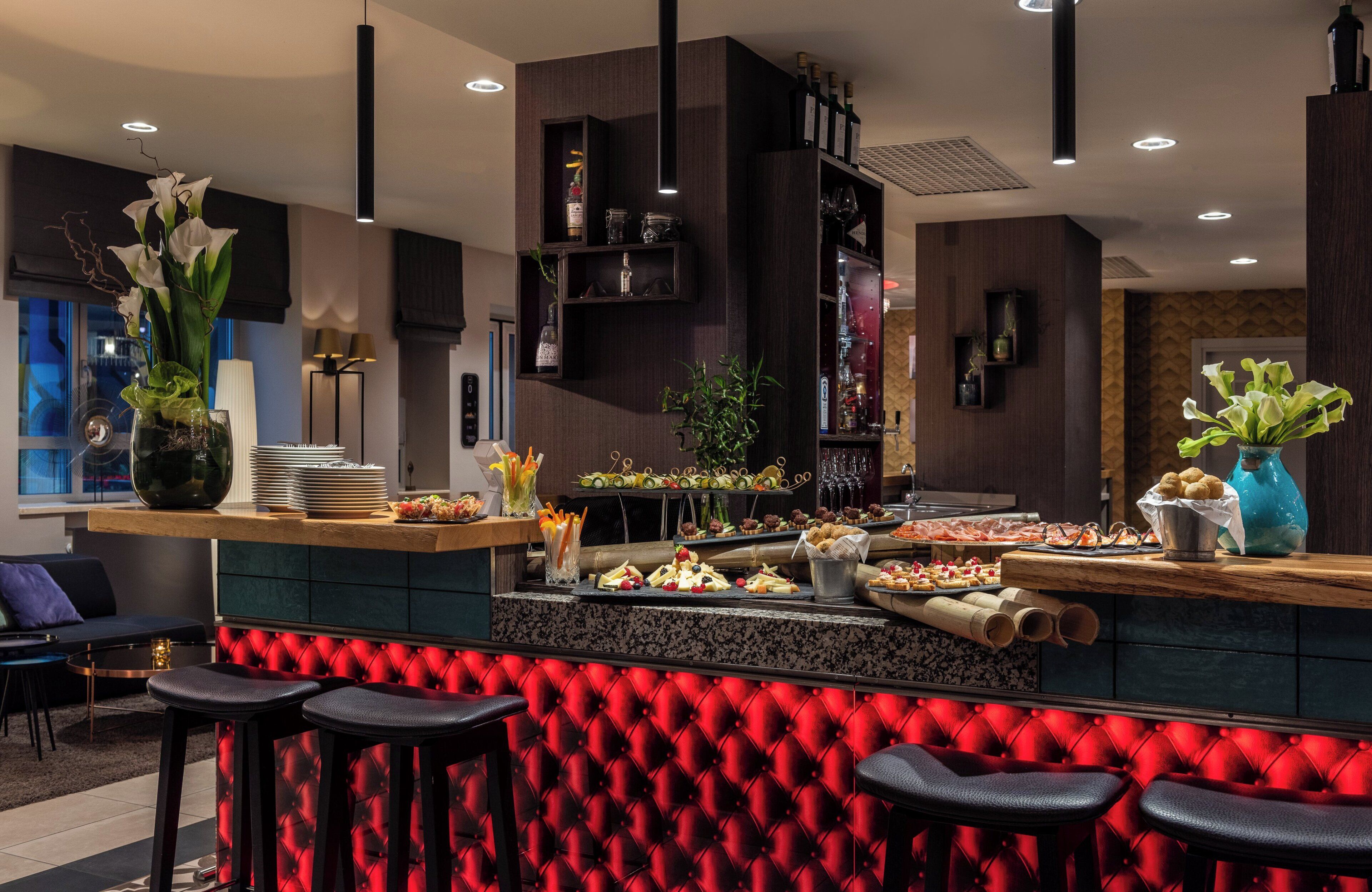 A modern bar area with a red cushioned bar stool seating, a variety of food and drink items on the counter, and a decorative vase with white flowers.