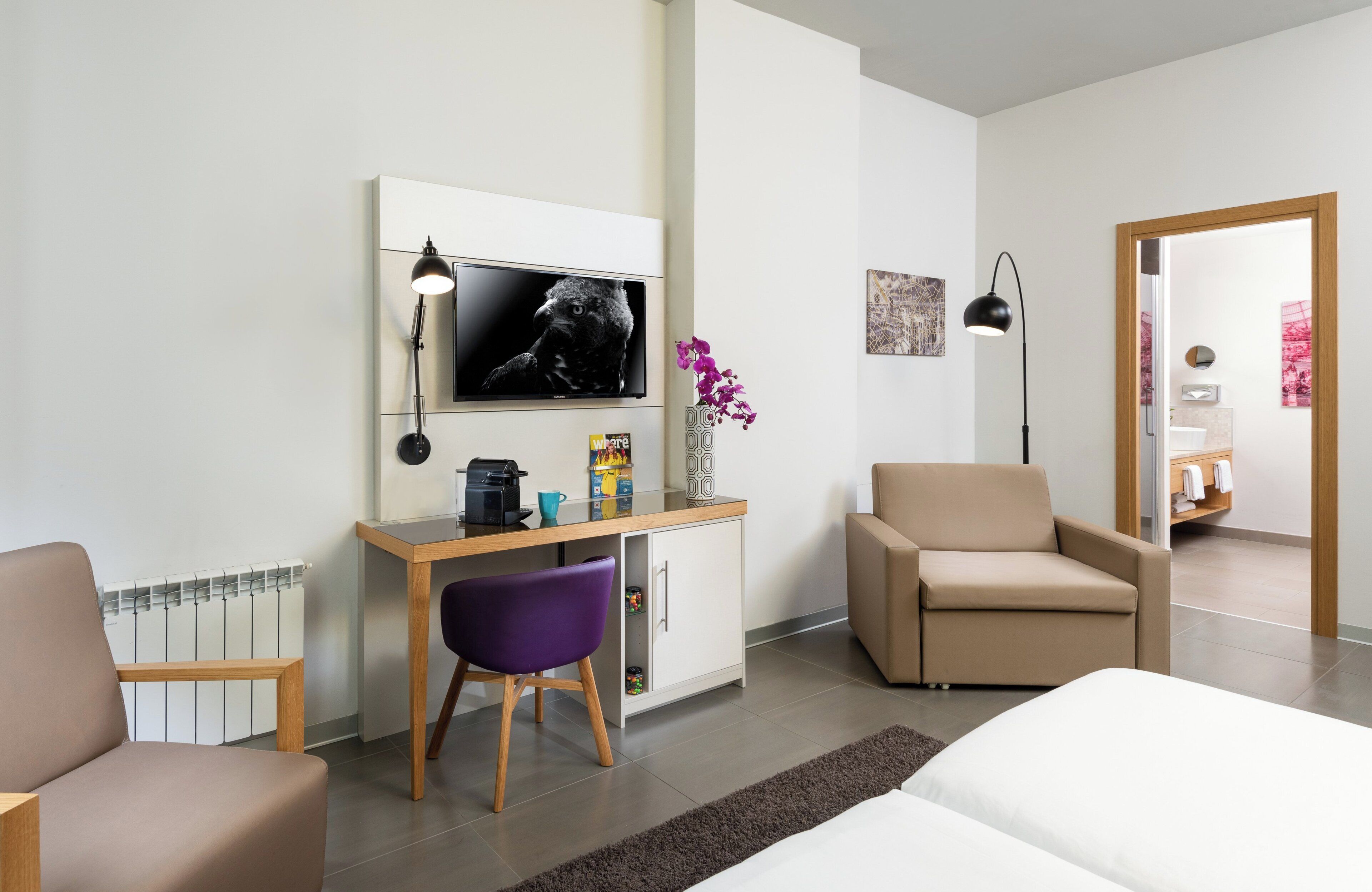 A modern hotel room with a bed, a desk with a computer, a chair, a radiator, and a TV.