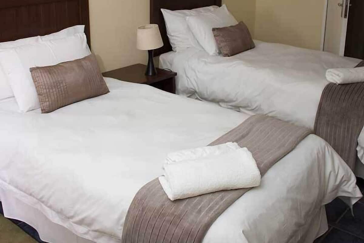 Ein Hotelzimmer mit zwei Betten, jeweils mit weißer Bettwäsche und braunen Kissen. Darüber hängt ein Gemälde, ein Nachttisch mit Lampe und eine Tür rechts.