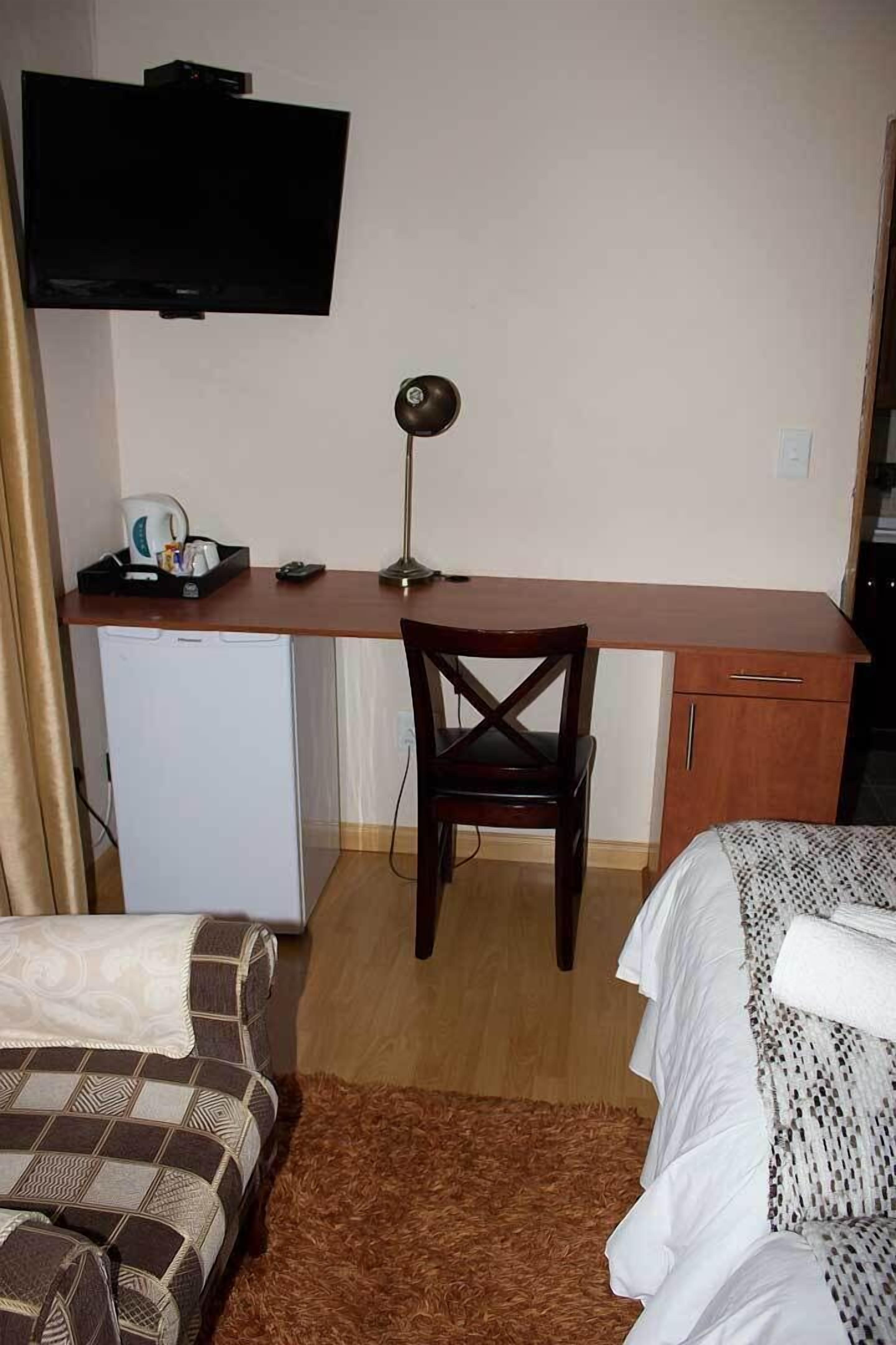 Habitación compacta con escritorio de madera, una silla, televisor montado sobre el escritorio, refrigerador pequeño y dos camas con ropa de cama estampada.