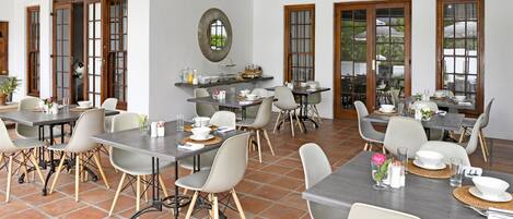 A spacious dining area with multiple tables and chairs, large windows, and a tiled floor.