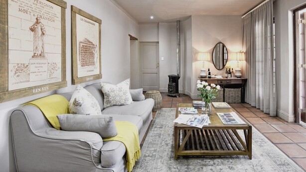 A living room with a gray sofa, yellow throw, and a coffee table with magazines. There are framed pictures on the wall and a lamp on the side table. - Mont d'Or Franschhoek (Franschhoek)