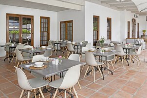 A spacious dining area with multiple tables and chairs, large windows, and a tiled floor.