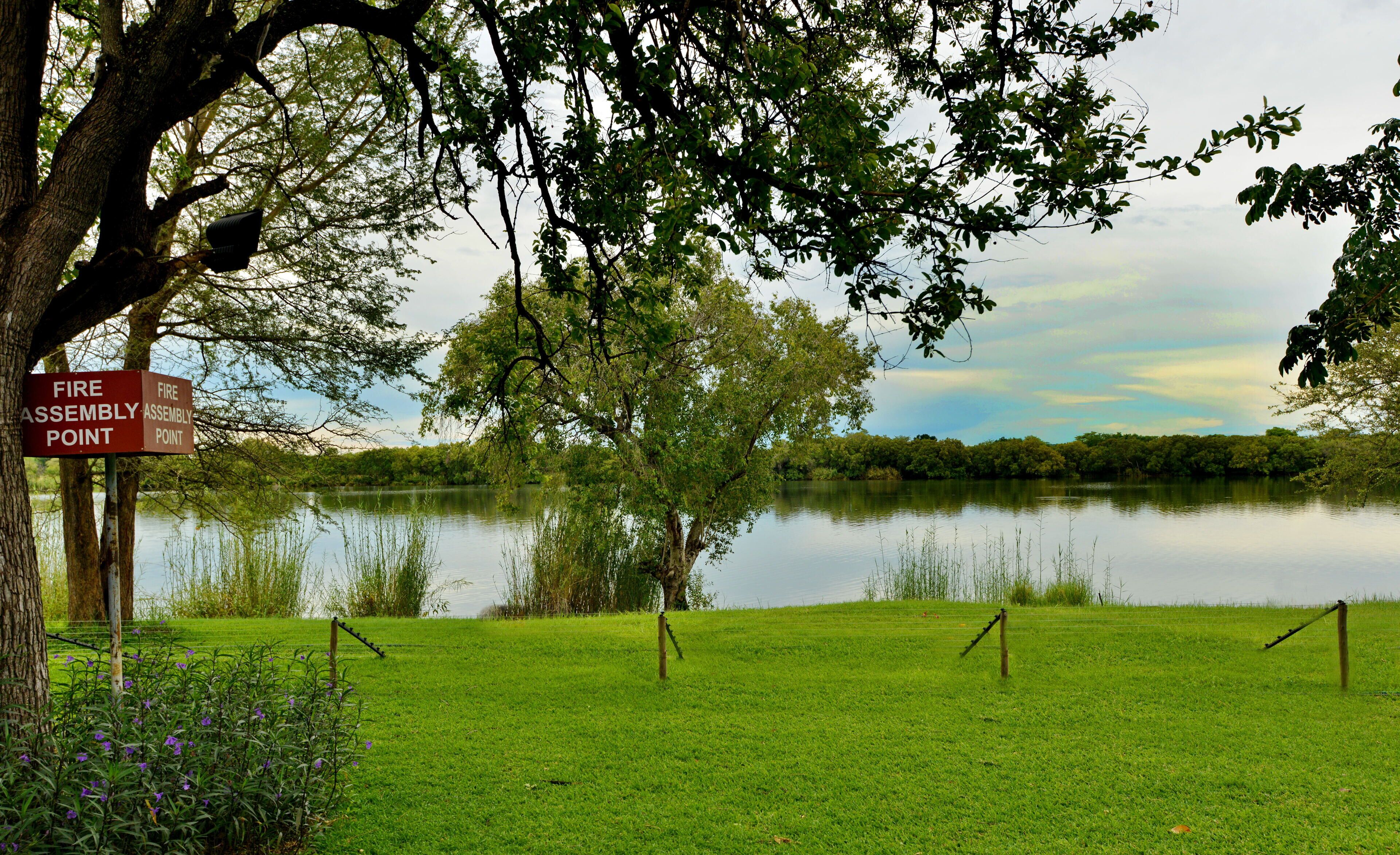A serene lakeside area with a fire assembly point sign, surrounded by lush greenery and a calm lake.