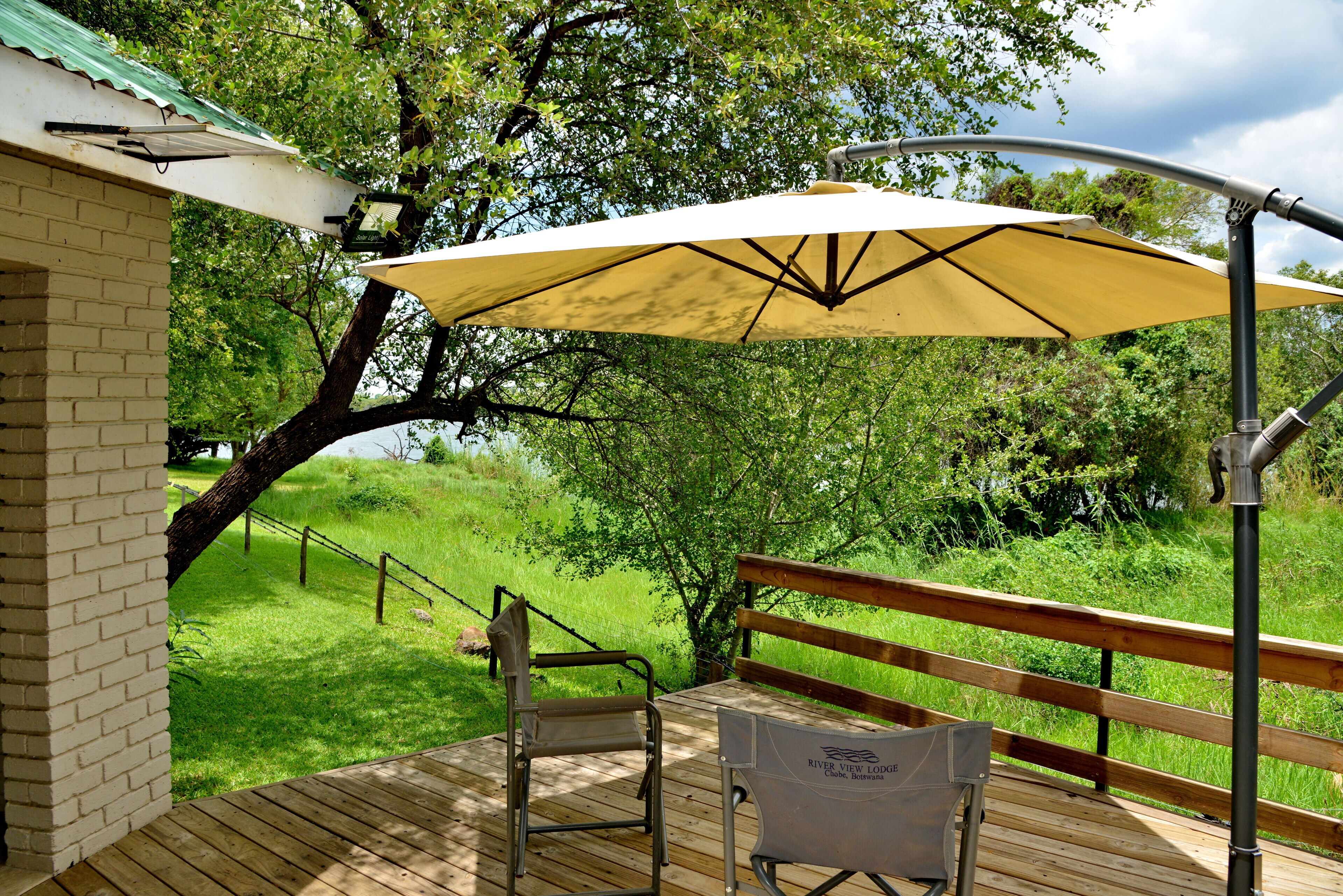 A wooden deck with a bench and a folding chair, a yellow umbrella, and a brick building in the background.