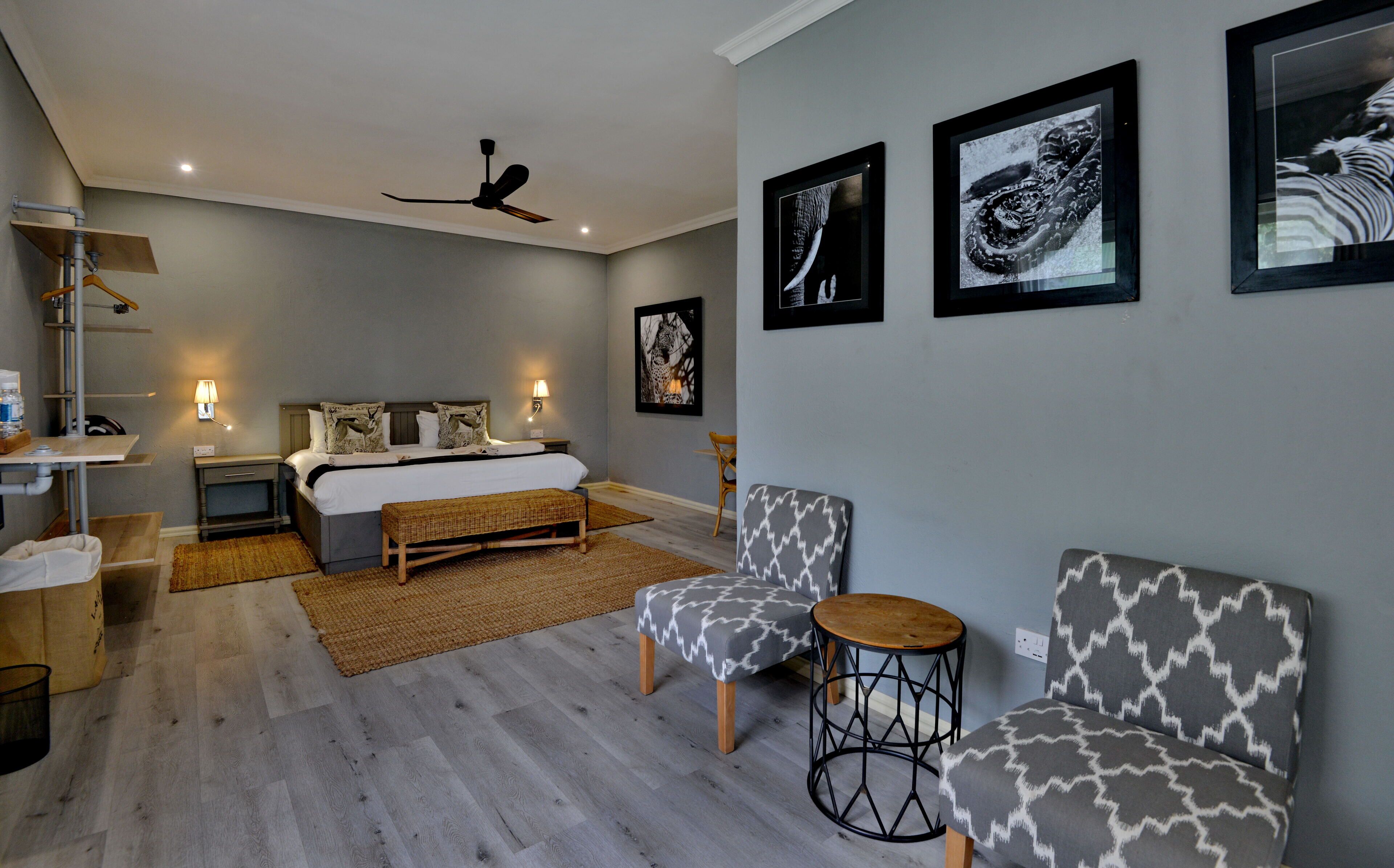 Een moderne slaapkamer met een bed, twee stoelen, een klein tafeltje, een plafondventilator en kunstwerken aan de muur.