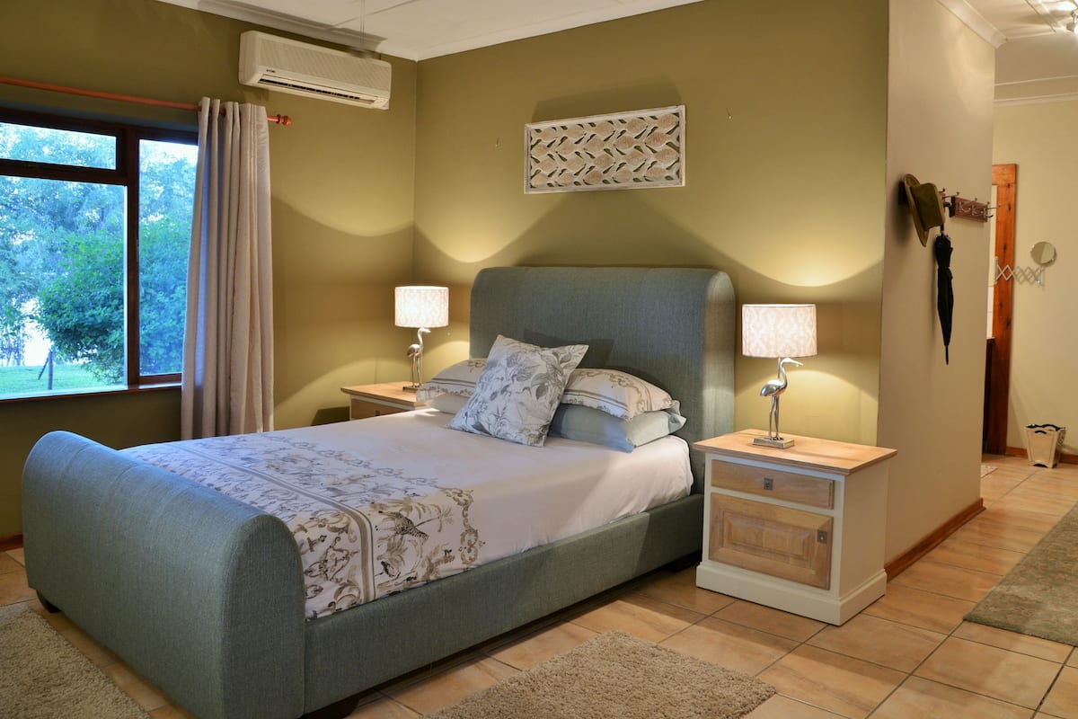 A bedroom with a bed, two bedside tables, a window with curtains, an air conditioning unit, and a decorative wall piece.