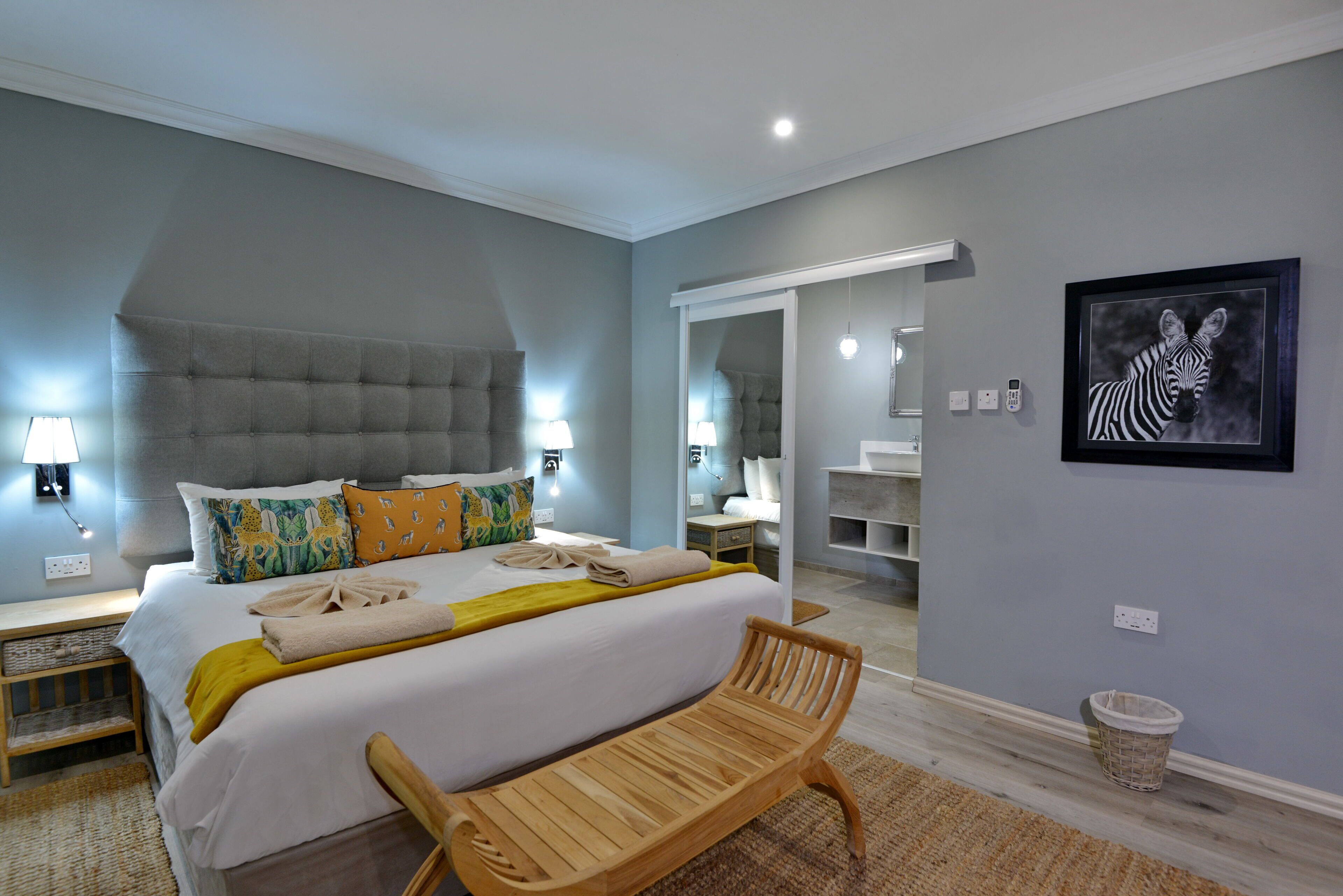 A modern bedroom with a large bed, bedside lamps, a wooden bench, a mirror, and a framed zebra artwork.