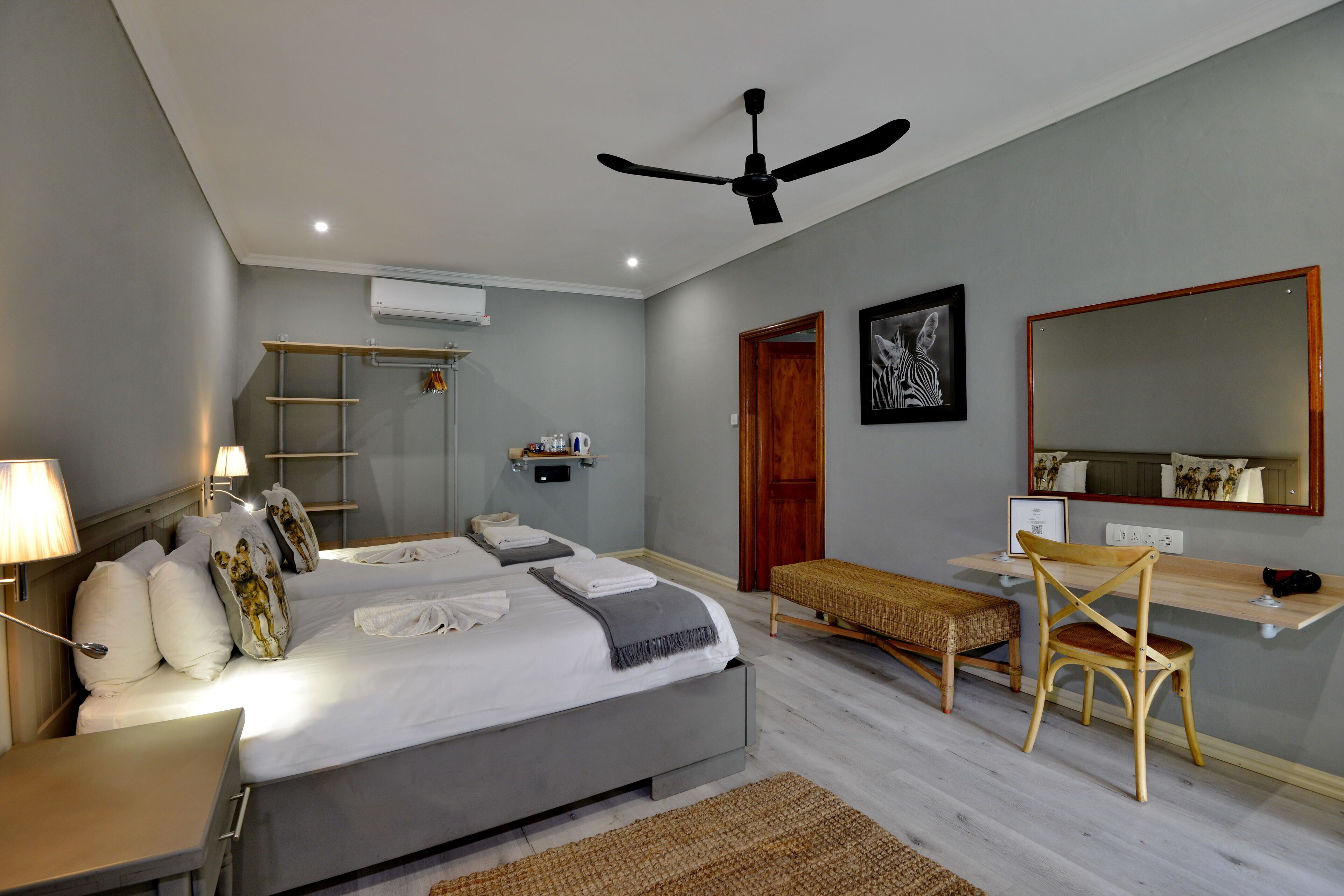 Un dormitorio con cama, mesitas de noche, escritorio, silla, ventilador de techo, espejo y estante en la pared.