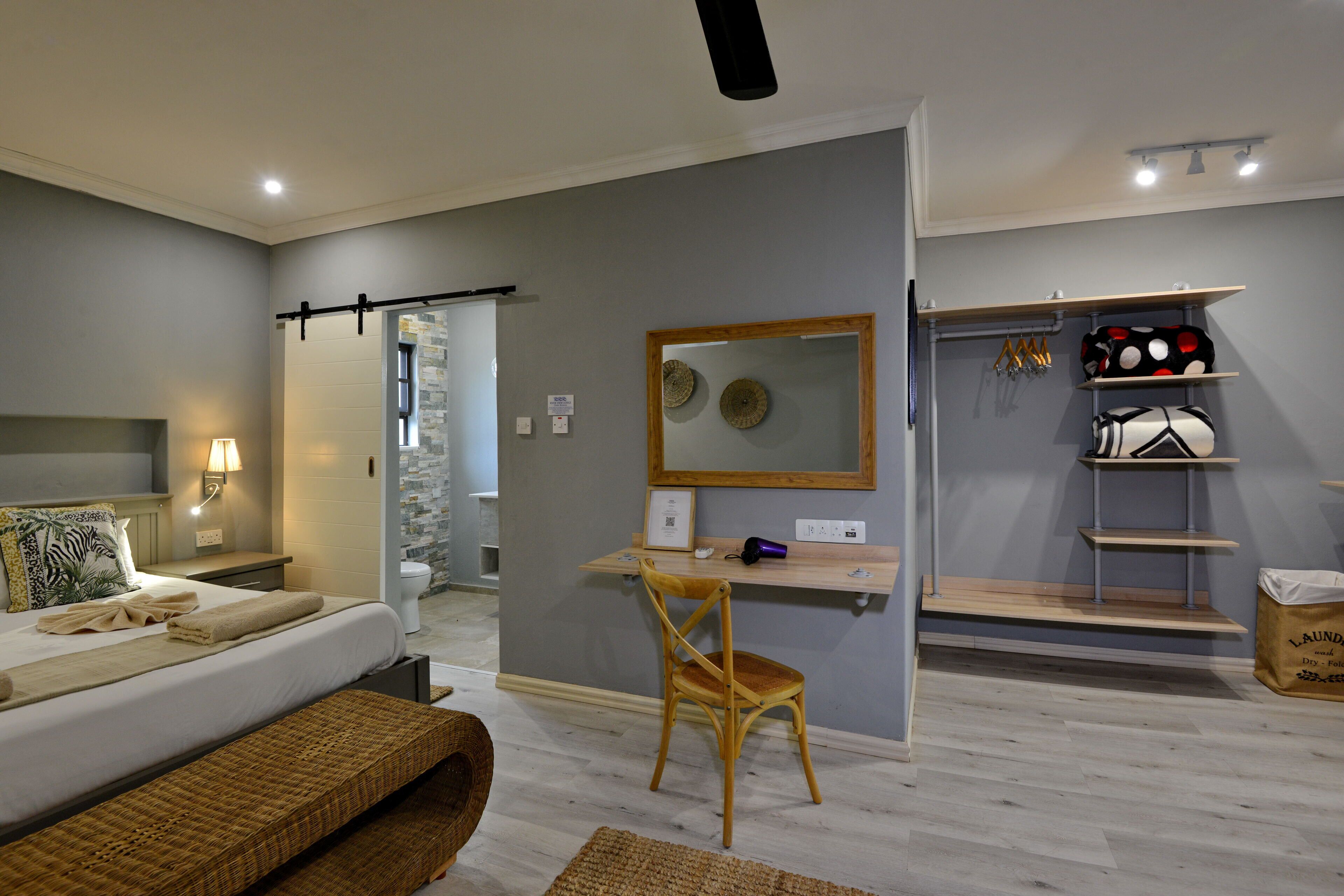 A modern bedroom with a bed, bedside table, lamp, wooden chair, desk, mirror, and shelves with towels and a laundry bag.