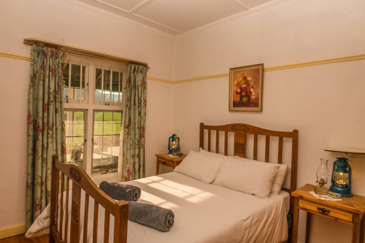 A bedroom with a wooden bed, a nightstand with a lantern, a window with floral curtains, and a painting on the wall.