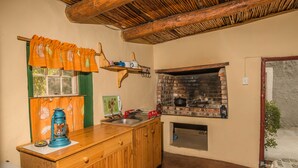 A rustic kitchen with wooden cabinets, a brick oven, and a window with orange curtains.