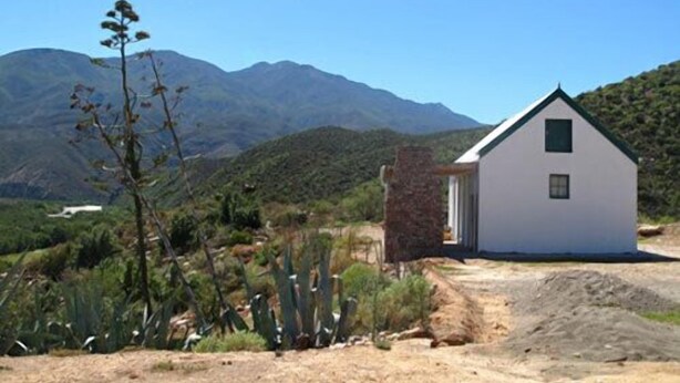 A small, white building with a stone chimney, surrounded by cacti and mountains in the background.