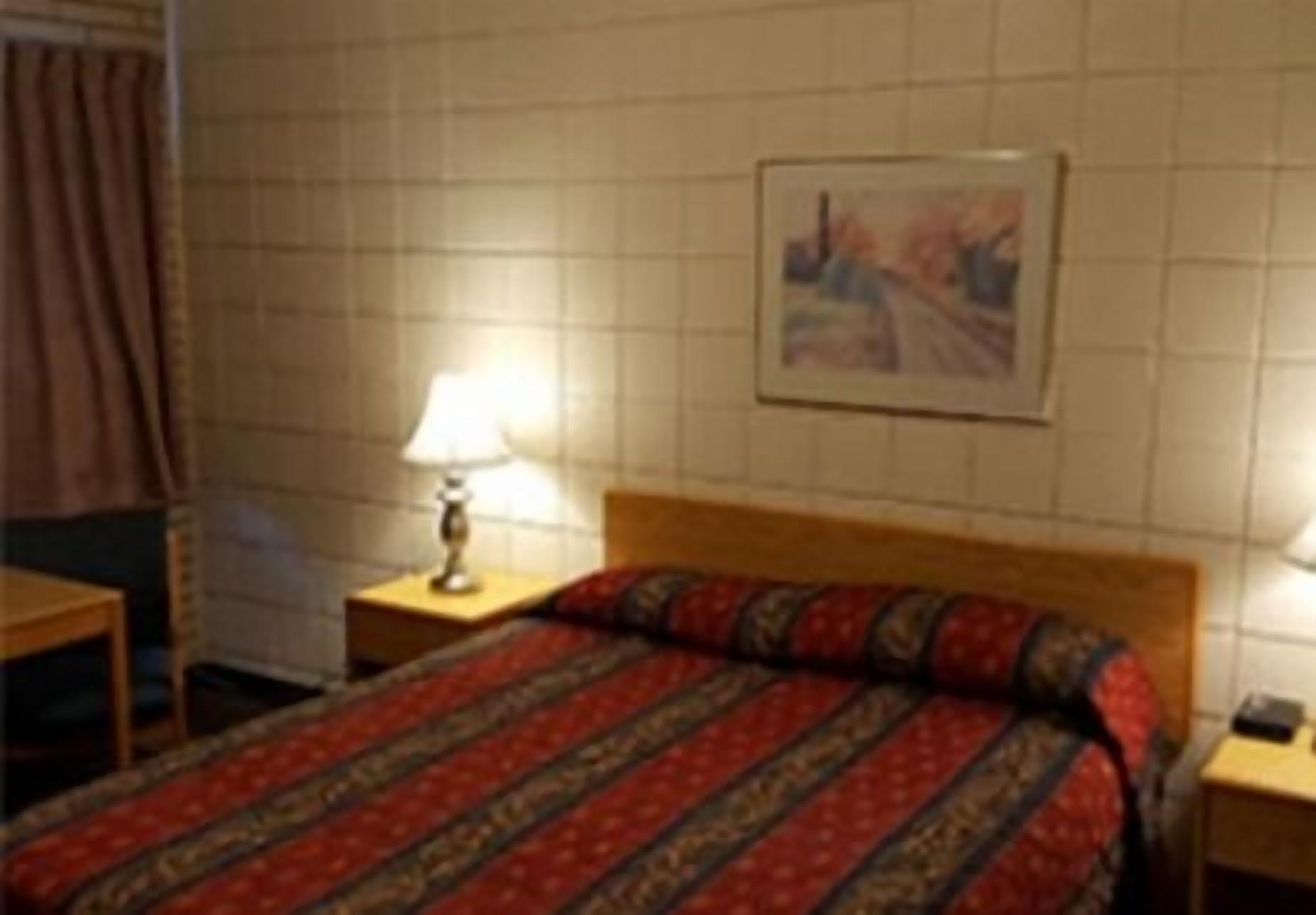 Une chambre d’hôtel avec un lit, une table de chevet avec une lampe, un tableau encadré au mur et une fenêtre avec des rideaux.