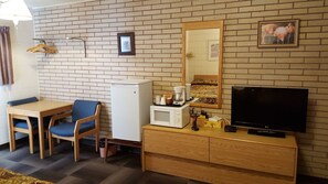 A compact hotel room with a brick wall, a small dining table with blue chairs, a microwave, a television, and a wooden cabinet.
