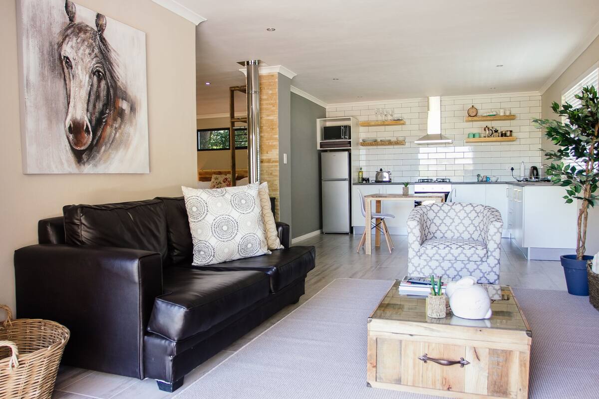 Un salon moderne avec un canapé en cuir noir, un fauteuil à motifs et une table basse en bois. Un grand tableau représentant un cheval orne le mur.