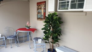 Pieni patio, jossa on kaksi metallituolia, punaisella kankaalla päällystetty pöytä, ruukkukasvi ja seinälle ripustettu taulu.