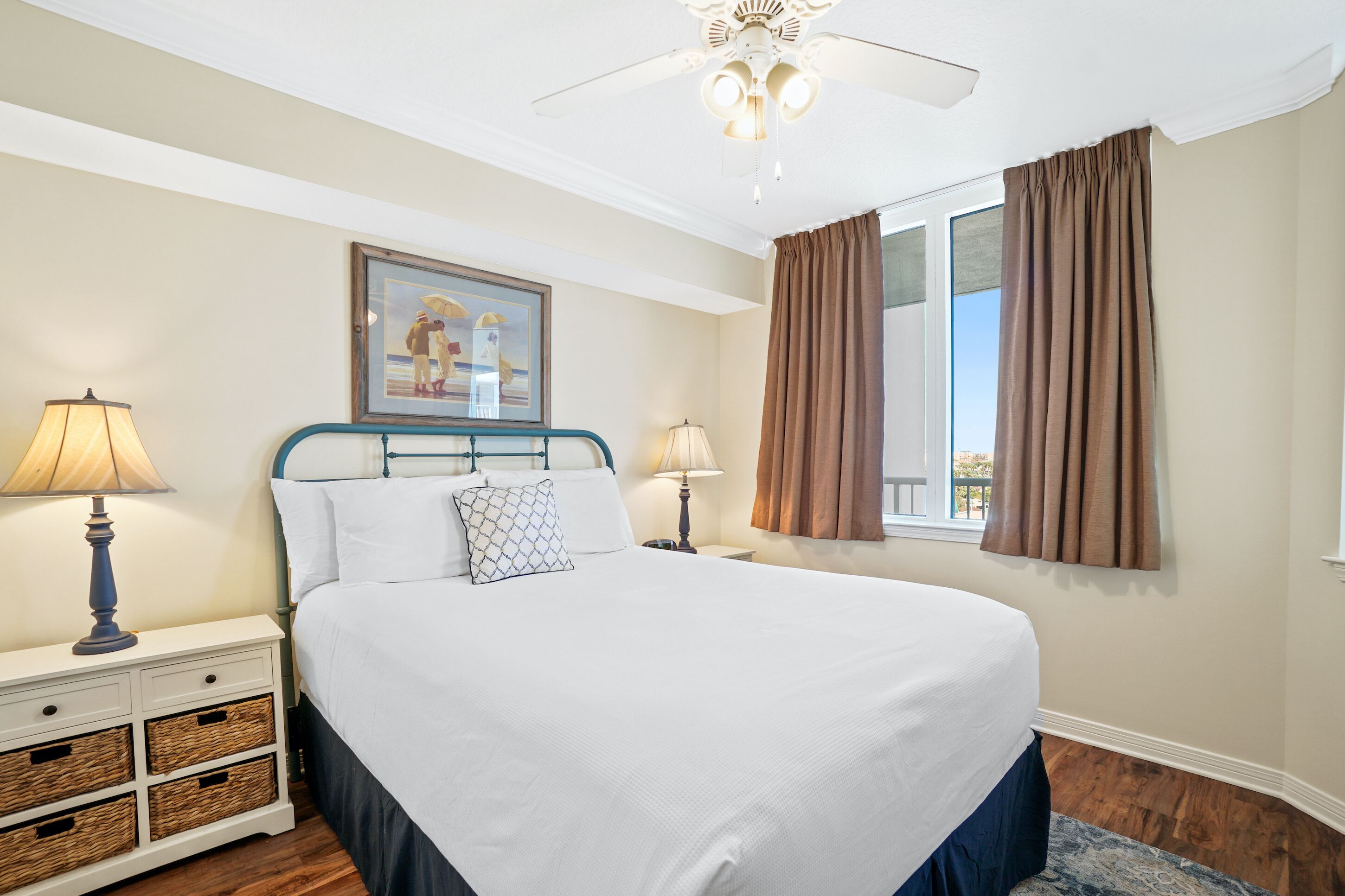 A neatly arranged bedroom with a large bed, two bedside tables with lamps, a framed picture on the wall, and a window with curtains.
