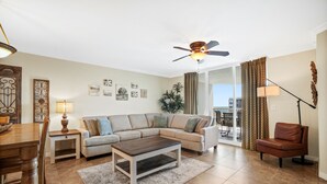 A living room with a sectional sofa, a coffee table, a dining table with chairs, a ceiling fan, and a view through a glass door. - Destin West Resort by Panhandle Getaways (Fort Walton Beach)