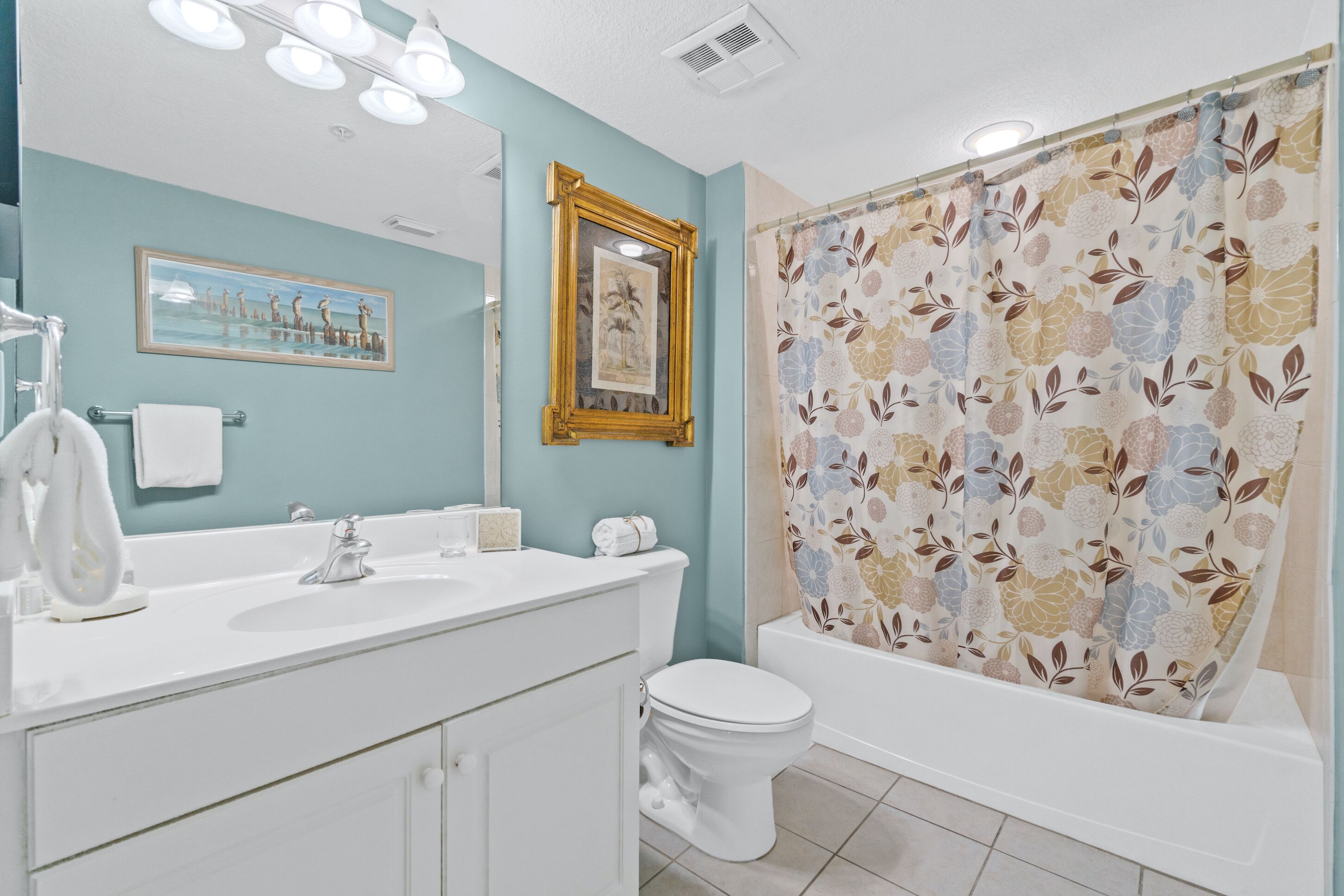 Un bagno con lavandino bianco, specchio e servizi igienici. Tenda da doccia con motivo floreale.