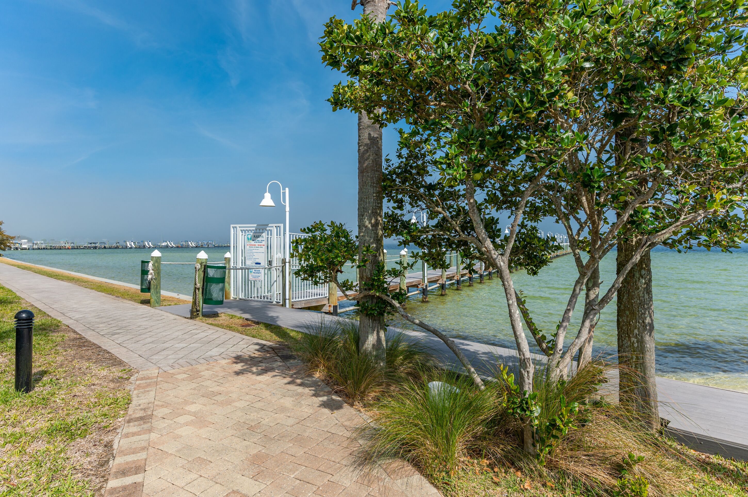 A waterfront walkway with a clear view of the water, a lamp post, and a trash receptacle.