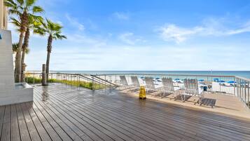 Een houten terras met lounge fauteuils en een waarschuwingsbord, met uitzicht op een strand met parasols.