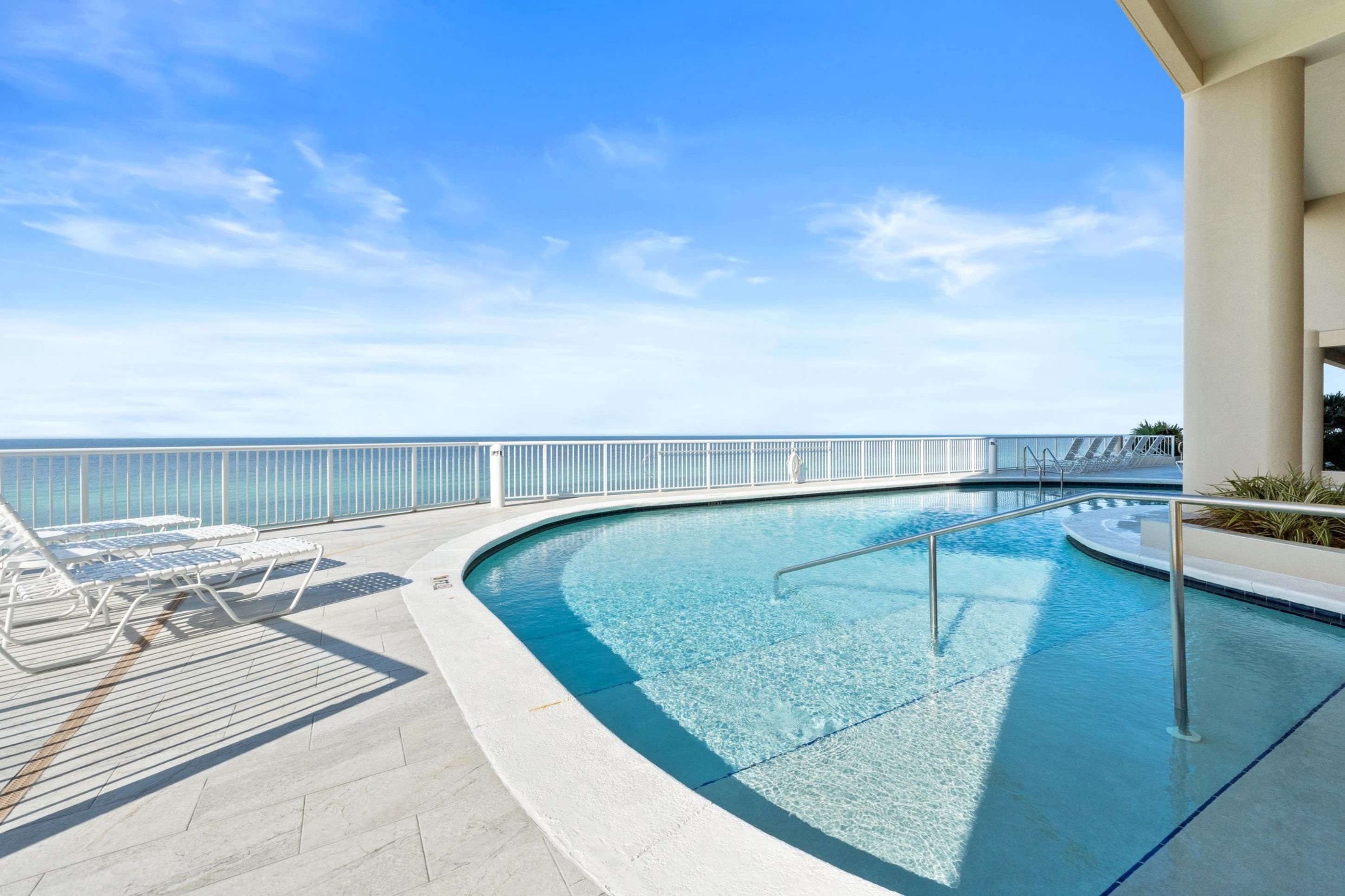 A pool area with a clear view of the ocean, lounge chairs, and a railing.